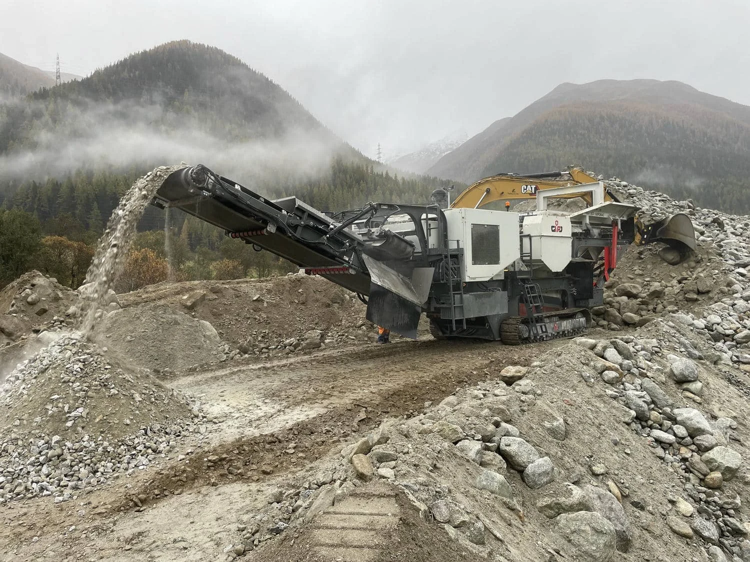 Mobiler Backenbrecher auf einer Baustelle im Gebirge, Förderband lädt gebrochenes Flussgestein auf einen Schüttkegel.