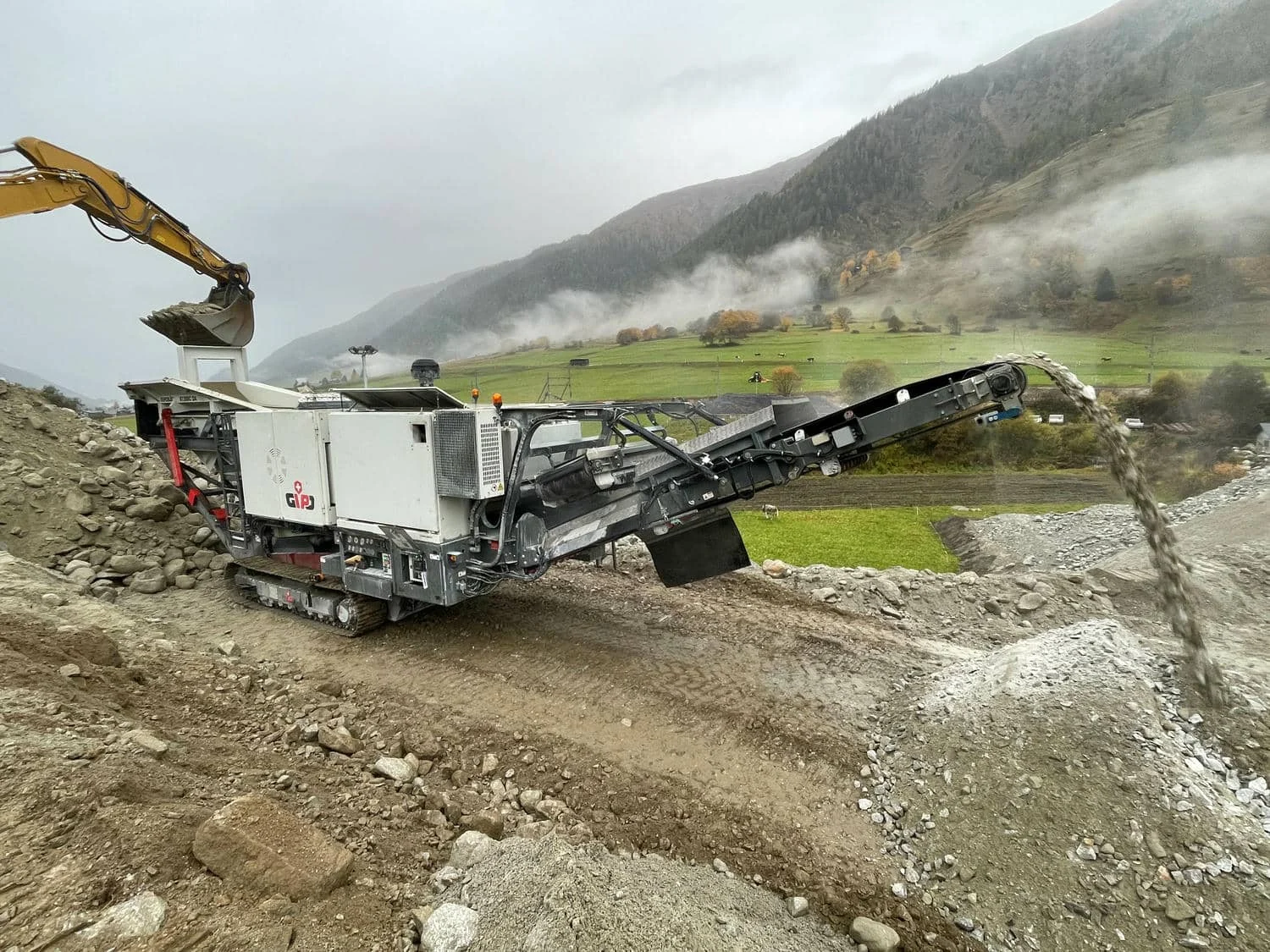 Bagger beschickt einen mobilen Brecher auf einer geneigten Kiesfläche, Berge und Tal im Hintergrund.