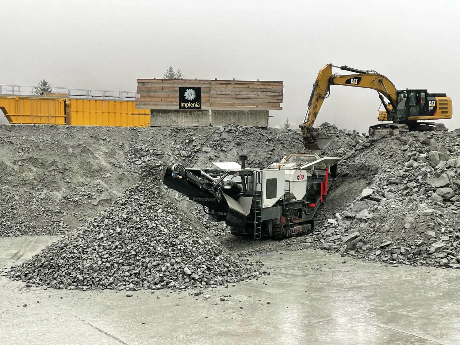Bagger lädt Bauschutt in einen mobilen Brecher, daneben ein großer Schüttkegel aus gebrochenem Material auf einer Betonfläche.