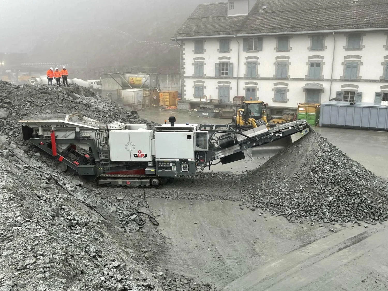 Mobiler Brecher verarbeitet Gesteinshaufen auf einem Industriegelände vor einem mehrstöckigen Gebäude, Mitarbeitende beobachten die Anlage.