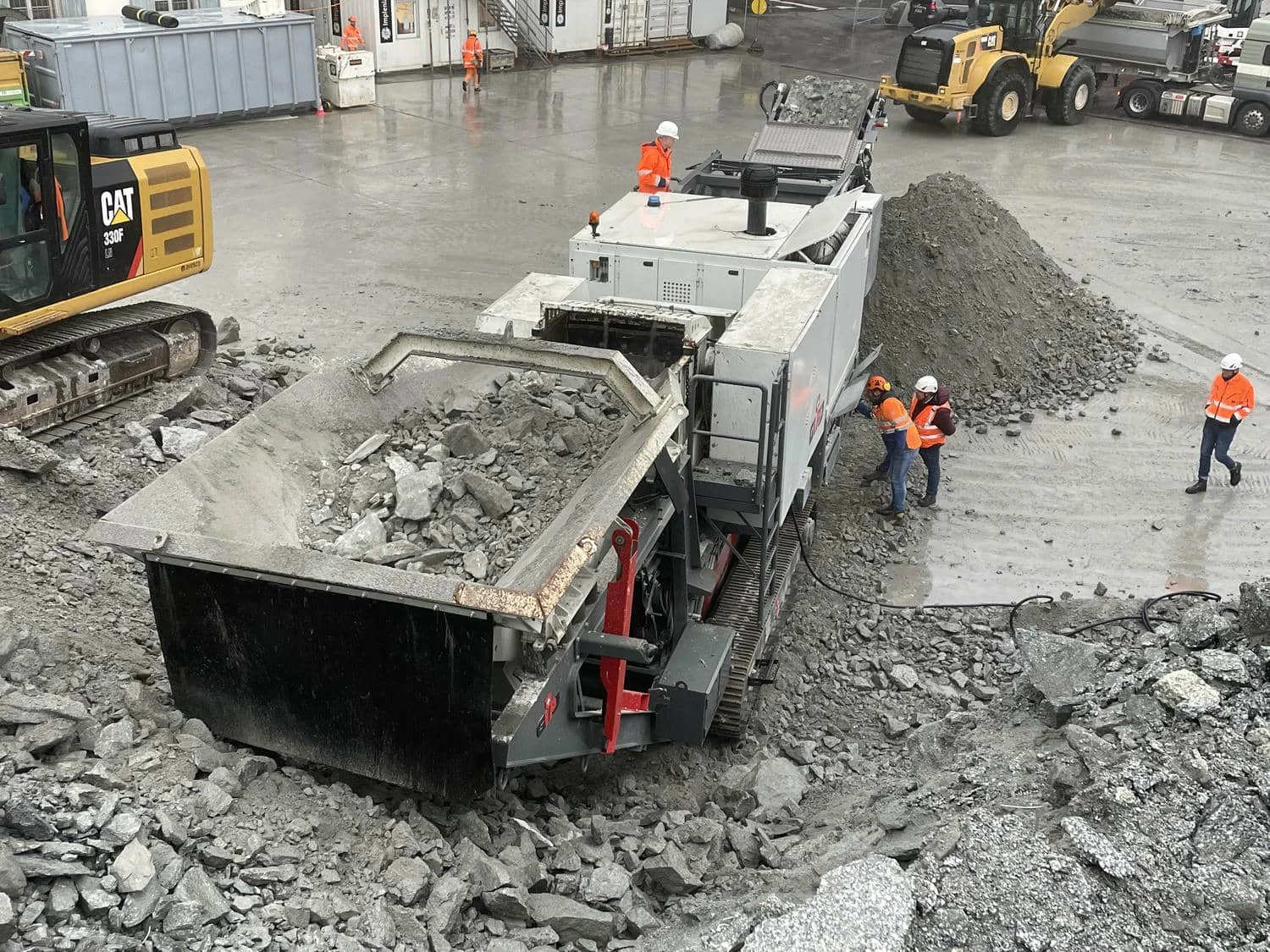 Nahaufnahme eines mobilen Brechers mit gefülltem Aufgabetrichter, mehrere Mitarbeitende in Warnkleidung arbeiten rund um die Maschine auf dem Hof.