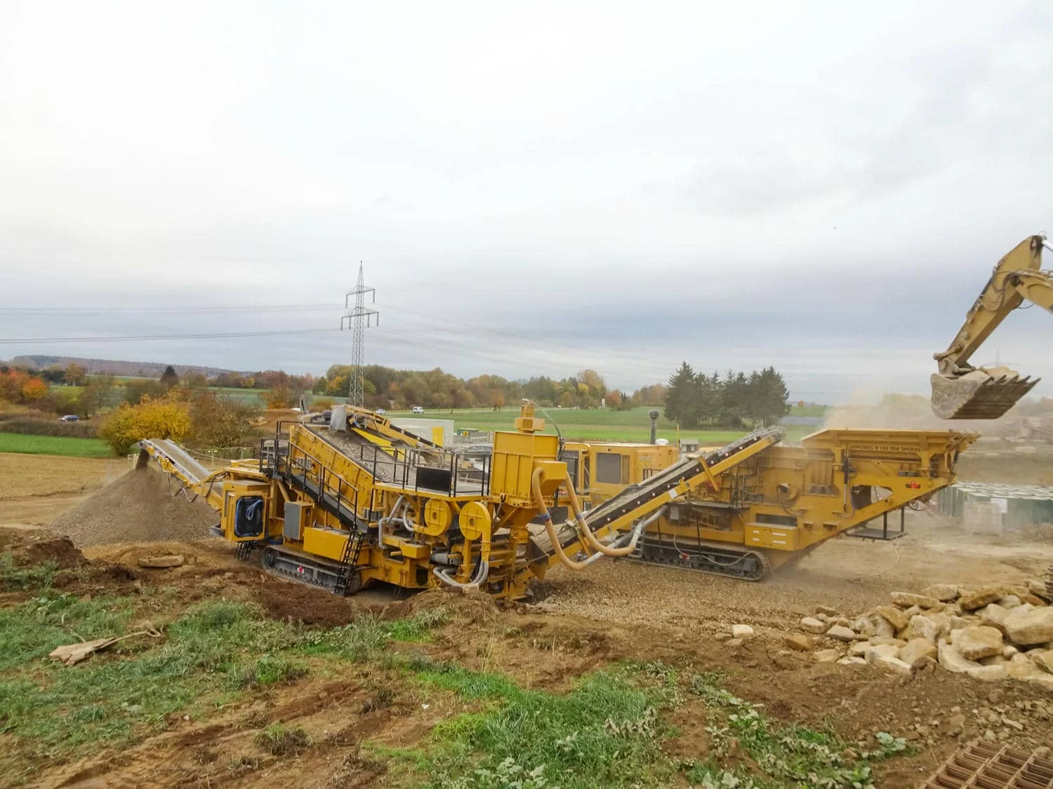 Mobile Brech- und Siebanlage im Einsatz auf unebenem Gelände, wobei ein Bagger große Steinblöcke in den Aufgabetrichter lädt.
